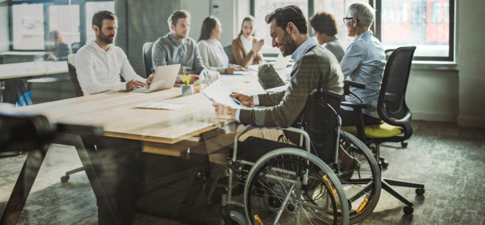 Happy male entrepreneur in a wheelchair going through paperwork during a meeting with his colleagues in the office. The view is through glass. Emploi des travailleurs handicapés : mettez-vous en conformité avec les obligations en vigueur depuis le 1er janvier 2020 !